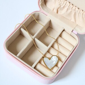 Peut inclure: Une boîte à bijoux rose avec une chaîne dorée et un pendentif en forme de cœur avec une incrustation de nacre blanche.