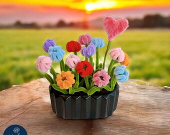 May include: A black ceramic planter filled with colorful, fuzzy flowers and a pink heart-shaped accent. The flowers are in shades of blue, pink, orange, red, and purple. The arrangement sits on a wooden surface with a sunset backdrop.