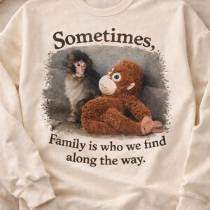 May include: Cream-colored sweatshirt featuring a graphic of a monkey and a stuffed monkey. The text reads "Sometimes, Family is who we find along the way." The sweatshirt has a crew neck and long sleeves.