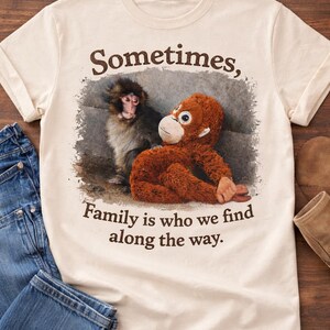 May include: Cream-colored t-shirt featuring a graphic of a monkey and an orangutan with the text "Sometimes, Family is who we find along the way." The shirt is paired with blue jeans and brown boots.