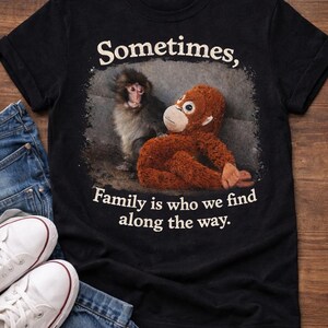 May include: Black t-shirt with the text "Sometimes, Family is who we find along the way." The shirt features a graphic of a monkey and a stuffed orangutan. White sneakers and blue jeans are visible.