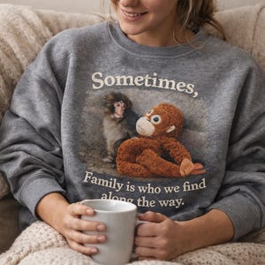 May include: A gray sweatshirt with the text "Sometimes, Family is who we find along the way." The sweatshirt features a graphic of two monkeys. A person is holding a white mug.