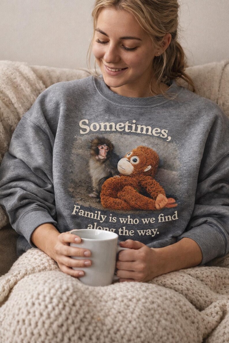 May include: A gray sweatshirt with the text "Sometimes, Family is who we find along the way." The sweatshirt features a graphic of two monkeys. A person is holding a white mug.