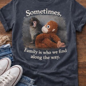 May include: Dark blue t-shirt with a graphic of two monkeys and the text "Sometimes, Family is who we find along the way." White sneakers and blue jeans are visible in the foreground. The shirt is made of a soft material.