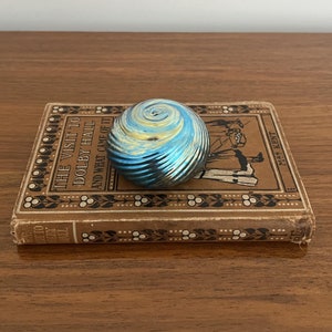 May include: A blue and gold iridescent glass paperweight with a swirling pattern sits on top of a vintage book titled "The Visit to Dolby Hall and What Came of It" by May Kent.