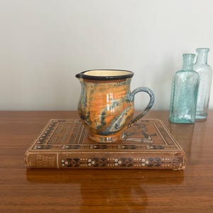 May include: A small, hand-painted ceramic jug with a black rim and handle. The jug has a blue, orange and yellow glaze with a swirling pattern. It is sitting on top of a vintage book.