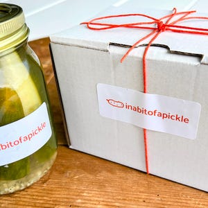 May include: A glass jar filled with green pickles featuring a gold lid and a white label with the red text inabitofapickle, alongside a white cardboard box tied with red string and a matching white label with the red text inabitofapickle.