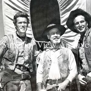 May include: Black and white photograph of three men in Western attire. They are wearing hats, vests, and belts. One man has a necklace. The background includes a covered wagon. The image is a vintage portrait.