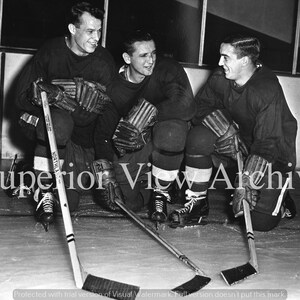May include: Black and white photograph of three hockey players kneeling on the ice. They wear dark jerseys, knee pads, and gloves. Hockey sticks are on the ice. The text "Superior View Archive" is across the image.