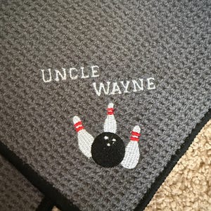 May include: Gray waffle-weave bowling towel with the embroidered text "UNCLE WAYNE" in white. A black bowling ball and three white bowling pins with red stripes are embroidered below the text. The towel has a black border and a metal clip.