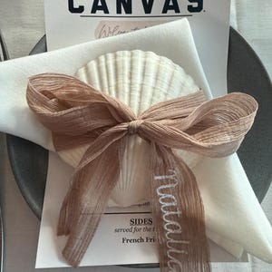 May include: A table setting featuring a white seashell, a white napkin, and a menu card. A light brown ribbon is tied around the seashell and napkin. The menu card displays the word "CANVAS" in bold black letters.