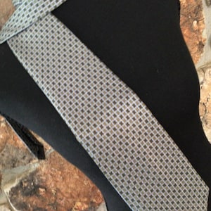 May include: A necktie featuring a repeating geometric pattern of small dark gray diamonds and light gray circles on a silver-toned background. The tie is displayed on a black mannequin form, showcasing the intricate woven texture and the pointed end.