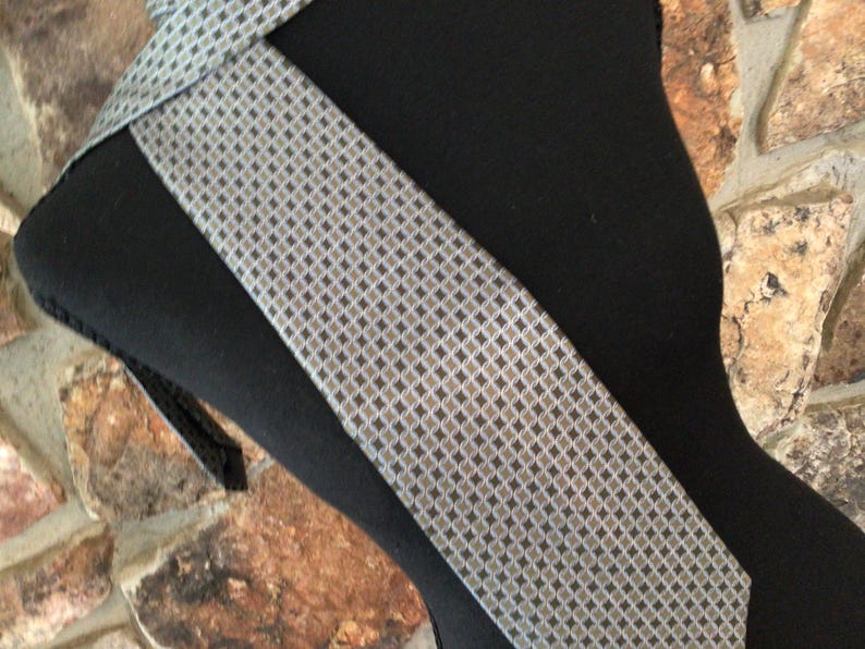 May include: A necktie featuring a repeating geometric pattern of small dark gray diamonds and light gray circles on a silver-toned background. The tie is displayed on a black mannequin form, showcasing the intricate woven texture and the pointed end.