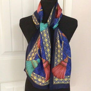 May include: A navy blue scarf featuring a repeating pattern of decorative tassels in shades of red, pink, and teal. The design includes gold-toned rope accents and a geometric border pattern in yellow and white, set against a deep blue background.