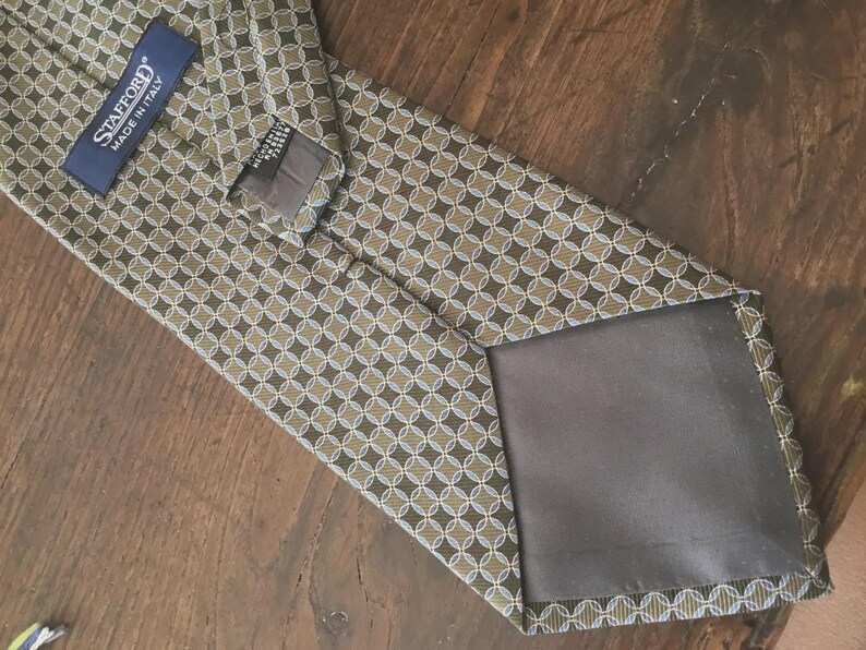 May include: A necktie featuring a repeating geometric pattern of interlocking circles in olive green, light blue, and white. The tie includes a dark blue label with the white text STAFFORD MADE IN ITALY and a smaller black care tag.