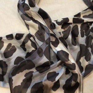 May include: A lightweight scarf with a black and gray animal print pattern. The scarf is made of a soft, flowing fabric and is draped in a casual style. The design features large, irregular shapes in varying shades of gray and black.
