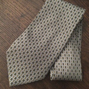 May include: A necktie featuring a repeating geometric pattern of interlocking circles and ovals in shades of olive green, tan, and white. The fabric has a subtle sheen, and the design is consistent across the entire surface of the folded necktie.