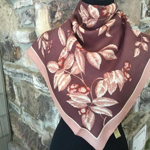 May include: A square scarf featuring a deep plum background with a repeating pattern of light pink and cream-colored floral branches and leaves. The scarf is finished with a solid light pink border along the edges, creating a distinct frame.