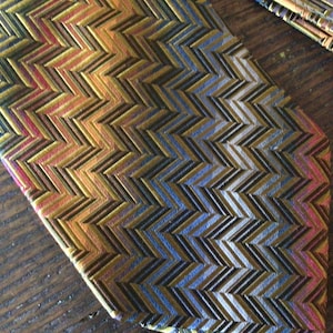 May include: Close-up of a patterned necktie with a zigzag design. The tie features a combination of colors, including gold, brown, blue, and red. The fabric has a textured appearance, and the pattern is repeated across the surface.