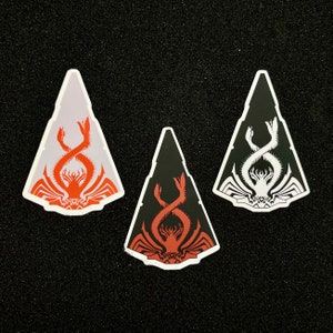 以下が含まれることがあります： 黒と白の生き物のデザインが施された三角形のステッカーが3枚。生き物は、カニまたはロブスターの様式化された抽象的な描写です。ステッカーは、黒と白、赤と白、白の3色のバリエーションがあります。