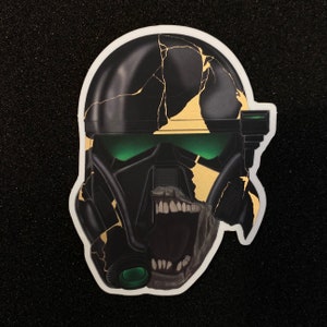 May include: A black and gold helmet with green visor and cracked paint. The helmet has a menacing expression with a wide open mouth and sharp teeth.