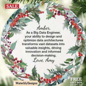 Peut inclure: Une décoration de Noël ronde avec une couronne florale à l'aquarelle. L'ornement porte le texte "Amber, As a Big Data Engineer...Love, Amy." Le mot "SALE" est dans un encadré rouge. L'ornement est suspendu à un sapin de Noël.