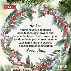 May include: A circular ceramic Christmas ornament featuring a watercolour floral wreath of green leaves and red berries. The ornament has text that reads "Amber, Your innovative solutions drive technology forward... Love, Amy." A "SALE" tag is visible.
