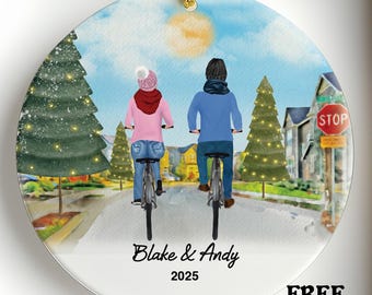 May include: A round ceramic ornament with a watercolor scene of two people riding bikes on a street lined with Christmas trees. The ornament has the text "Blake & Andy 2025". A red "SALE" banner is at the top.