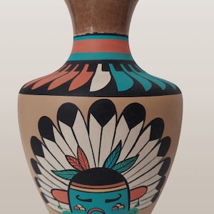 May include: A tan ceramic vase with a Native American design. The vase has a brown top, orange and turquoise bands, and a painted image of a face with a headdress.