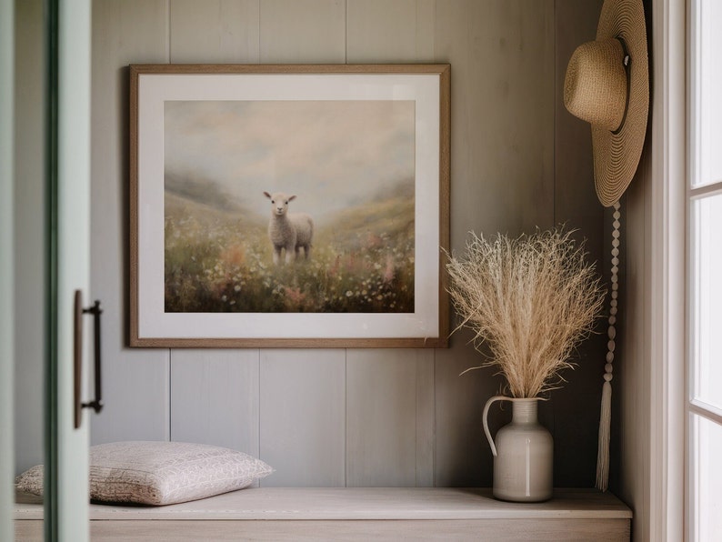 May include: A framed artwork featuring a lamb standing in a field of wildflowers. The artwork is in a light wood frame and hangs on a gray wall. A straw hat and a vase with dried grasses are also visible.