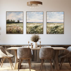 May include: Three framed landscape paintings depicting a field of white wildflowers with a blue sky and fluffy clouds in the background. The paintings are hung on a white wall above a wooden dining table with four chairs.