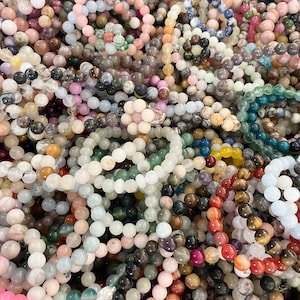 May include: A large pile of colorful gemstone bracelets. The bracelets are made of various types of stones, including amethyst, rose quartz, and tiger's eye.