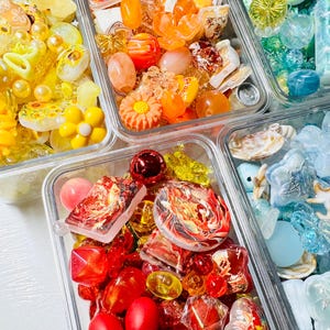 May include: Four clear rectangular containers filled with assorted craft beads and charms. One container holds yellow sunflower and floral beads, another orange and peach-toned pieces, a third blue sea-themed items, and the fourth red marbled beads.