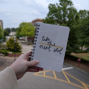 Puede incluir: Una libreta blanca de espiral con las palabras "take the next shot" escritas en tinta negra.