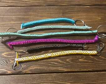 May include: Five colorful keychains made of woven plastic beads. The keychains are in shades of blue, green, purple, black, and yellow. Each keychain has a metal ring or clasp attached.