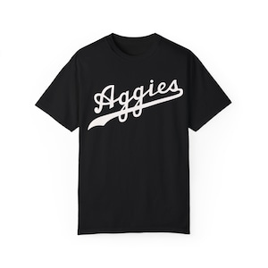 May include: Black t-shirt with the word "Aggies" in white script across the chest. The word has a curved underline. The shirt has short sleeves and a crew neck.