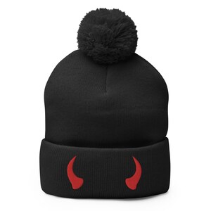 May include: A black beanie with a large black pom-pom on top. The beanie features embroidered red devil horns near the brim. This winter hat is a fun accessory.