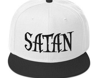 Puede incluir: Gorra de béisbol blanca con visera negra y botón negro. La palabra "SATAN" está impresa en letras negras grandes y estilizadas en la parte delantera de la gorra.