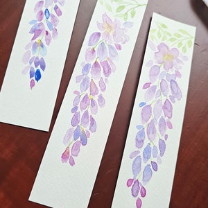 Op de afbeelding: Drie aquarel-bladwijzers met paarse en blauwe blauwe regen bloemen met groene bladeren. De bladwijzers zijn geschilderd op wit papier.