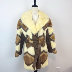 May include: A vintage coat with a cream-colored fur collar and geometric patchwork design. The coat features brown leather accents, a cream-colored fur body, and a matching belt. The coat is displayed on a mannequin.