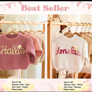 Op de afbeelding: Twee baby truien op houten hangers. Eén is roze met de naam "Hallie" in crèmekleurig garen en een witte bloem. De andere is wit met de naam "Amelia" in roze garen en een witte bloem. Beide zijn maat 0-3M.