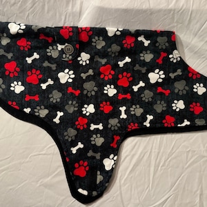 Könnte beinhalten: Ein Hundemantel mit schwarzem Hintergrund und einem Muster aus roten, weißen und grauen Pfotenabdrücken und Knochen. Der Mantel hat einen Knopfverschluss und eine schwarze Einfassung.