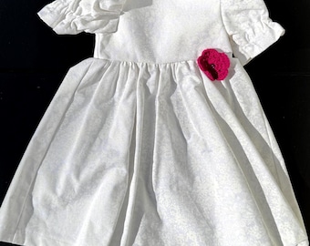 Puede incluir: Un vestido blanco con mangas cortas abullonadas y una falda fruncida. El vestido presenta una pequeña flor rosa brillante en el corpiño. La tela tiene un sutil estampado floral. El vestido está diseñado para un niño.