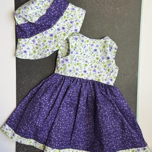 May include: A sleeveless dress and matching sun hat set featuring a white fabric with a small purple and green floral print on the bodice and hat crown, contrasted with a solid purple fabric with a darker purple floral pattern on the skirt.