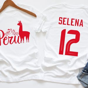 May include: Two white t-shirts with red designs. One shirt features a llama, mountains, and the word "Peru". The other shirt has the name "Selena" and the number "12" printed on the back.