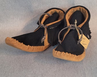 May include: A pair of black and brown moccasins with fringe. The moccasins are tied with a brown leather lace. The tag says "Black + Brown".