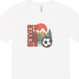 May include: White t-shirt with a graphic featuring a soccer ball, mountains, trees, and a setting sun. The text "DENVER SUMMIT FC" is printed vertically in orange and brown.