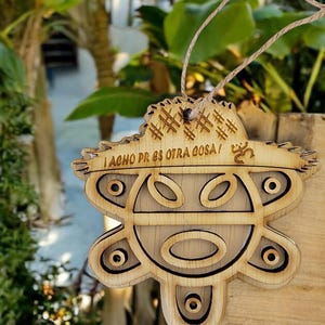 May include: Wooden ornament with a carved design and the text "¡ACHO PR ES OTRA COSA!". The ornament has a natural wood color and a string for hanging. The design includes geometric shapes and a face.