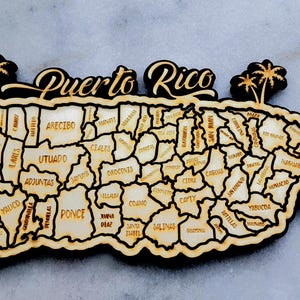 Op de afbeelding: Houten kaart van Puerto Rico met de omtrek van het eiland en de interne gemeenten. De woorden "Puerto Rico" staan bovenaan, met accenten van palmbomen en bloemen. De kaart is lichtbruin met een zwarte rand.