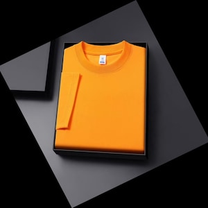 以下が含まれることがあります： 黒い箱に入った、折りたたまれた鮮やかなオレンジ色のTシャツ。 Tシャツの袖が見え、折りたたまれた衣服から伸びています。 シャツは柔らかく快適な素材で作られているようです。